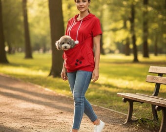 Puede incluir: Una sudadera con capucha roja de manga corta que presenta un gran bolsillo frontal tipo canguro con un pequeño perro gris asomándose por la abertura. La sudadera tiene cordones blancos y un bordado de huellas negras en el bolsillo, con vaqueros azules.