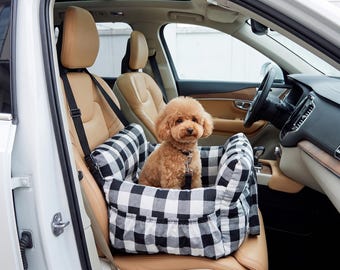 Puede incluir: Un asiento elevador para mascotas con estampado de cuadros vichy en blanco y negro, interior acolchado y correas negras ajustables. Un perro pequeño de color marrón claro y pelo rizado está sentado dentro, sujeto por una correa corta negra.