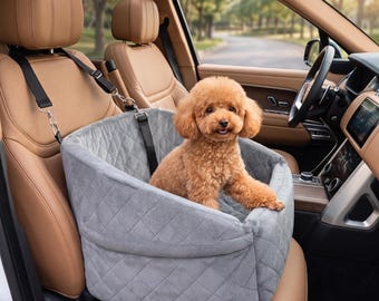 Puede incluir: Un asiento elevador para mascotas de color gris claro acolchado con correas negras ajustables aseguradas a los reposacabezas de un asiento de coche de cuero color canela. Un perro pequeño, esponjoso y de color marrón claro está sentado dentro.