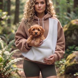 May include: A tan and white color-blocked hooded sweatshirt with a large front pouch pocket, worn with olive green athletic shorts. The sweatshirt features a drawstring hood and long sleeves, providing a casual look for outdoor activities.