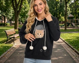 Puede incluir: Una sudadera negra de manga larga con capucha que presenta un bolsillo frontal con una ventana de malla que contiene un gatito pequeño, esponjoso y de color naranja claro. Tiene cordones blancos con pompones y un gráfico de huella.