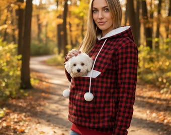 Puede incluir: Una sudadera con capucha de cuadros rojos y negros que presenta un bolsillo frontal grande diseñado para llevar un perro pequeño. La prenda incluye un forro de capucha blanco, cordones blancos largos con pompones blancos y un dobladillo rojo.