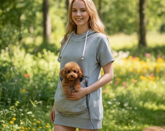 Puede incluir: Un vestido tipo sudadera con capucha de color gris claro y manga corta que presenta un bolsillo frontal grande. Un perro pequeño de color marrón y pelo rizado está colocado dentro del bolsillo. La prenda incluye cordones blancos y cremallera.