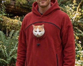 May include: A rust-colored long-sleeve hooded sweatshirt featuring a circular chest pouch with a drawstring closure. A white and orange cat is visible peering out from the pouch. The garment includes black zippered accents and ribbed cuffs.