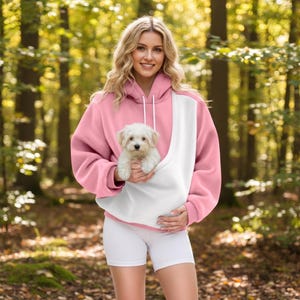 May include: A pink and white hooded sweatshirt featuring a large front pouch pocket designed to carry a small white puppy. The hoodie has long sleeves, a drawstring hood, and a color-blocked design with a white center panel and pink sides.