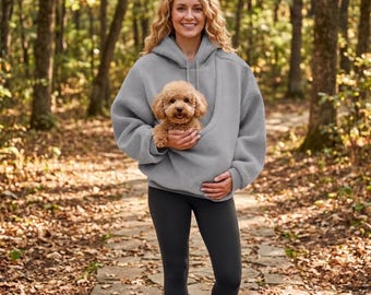 May include: A light gray fleece hoodie featuring a large front pouch pocket designed to carry a small dog. The hoodie has long sleeves, a hood with matching drawstrings, and a soft, textured fabric finish. A small brown poodle is inside the pouch.