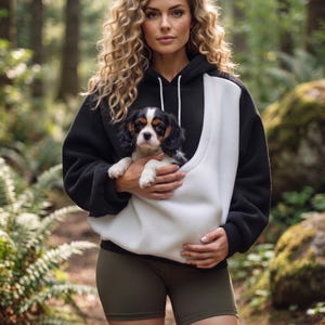 May include: A black and white hooded sweatshirt featuring a large front pouch pocket designed for carrying a small dog. The hoodie has long sleeves, a drawstring hood, and a solid black body with a contrasting white panel on the front.