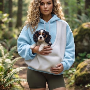 May include: A light blue and white fleece hoodie featuring a large front pouch pocket designed for carrying a small dog. The hoodie has long sleeves, a hood with white drawstrings, and a soft textured finish, paired with olive green shorts.