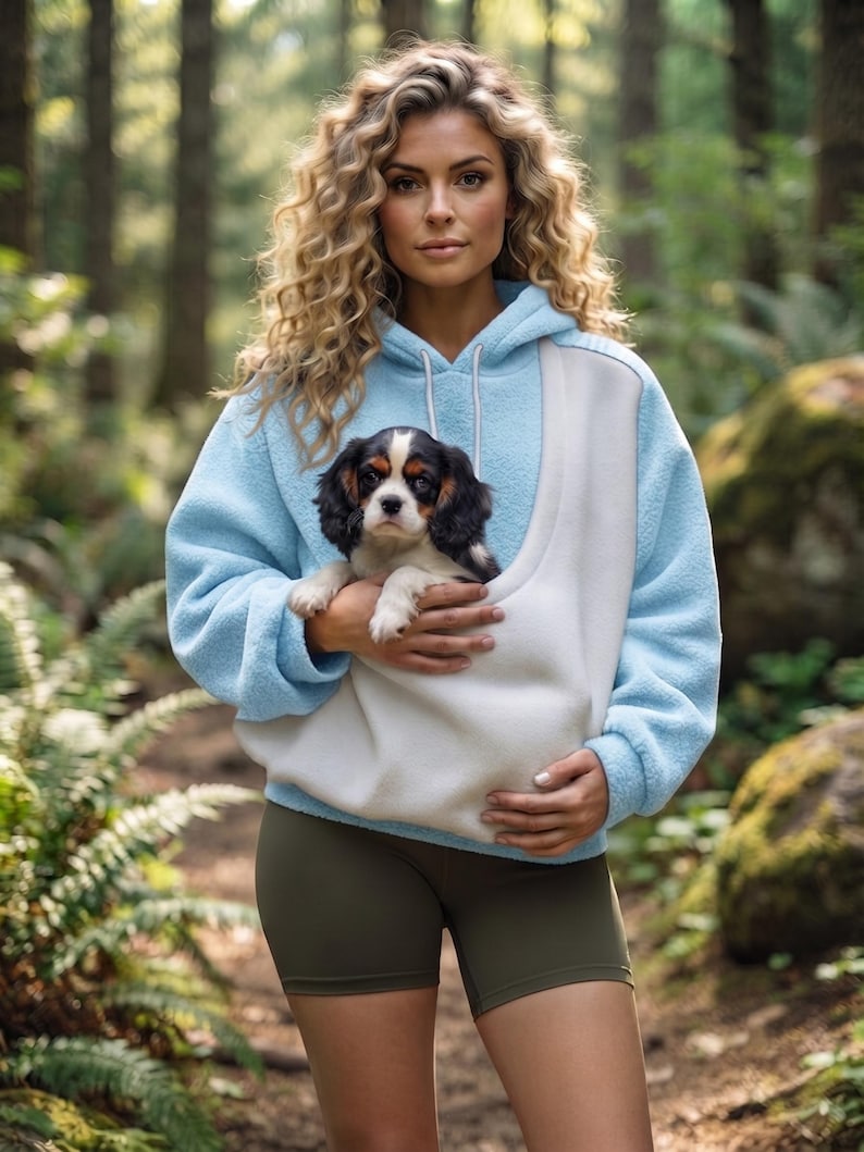 May include: A light blue and white fleece hoodie featuring a large front pouch pocket designed for carrying a small dog. The hoodie has long sleeves, a hood with white drawstrings, and a soft textured finish, paired with olive green shorts.