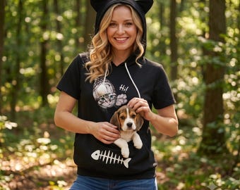 Puede incluir: Una sudadera negra de manga corta con orejas de gato en la capucha y cordones blancos. El frente muestra un gráfico de un gato con gorro de punto, el texto "Hi!" y un esqueleto de pescado blanco sobre un bolsillo tipo canguro.