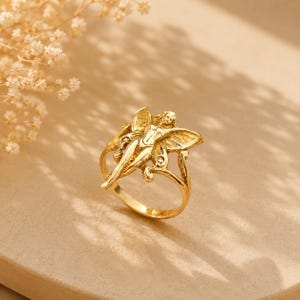 May include: A gold-toned ring featuring a detailed, sculpted figure with outstretched wings. The central design depicts a winged being with a slender form, integrated into a smooth, circular band. The metal has a polished, reflective finish.