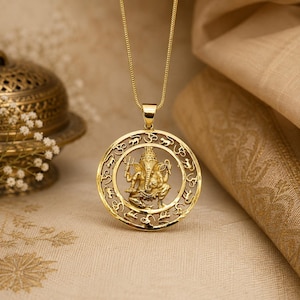 May include: A gold-toned pendant necklace featuring a circular frame with intricate cut-out details of the deity Ganesha in the center, surrounded by repeating Om symbols and small animal motifs, suspended from a delicate matching gold-toned chain.