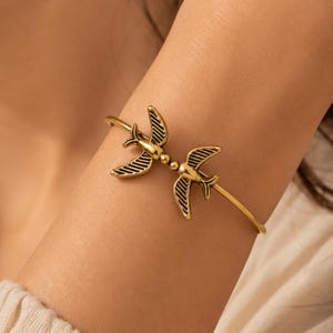 May include: A gold-toned metal cuff bracelet featuring two stylized bird motifs facing away from each other. The birds have detailed wing patterns with dark recessed lines and are connected at the center by a small spherical bead element.