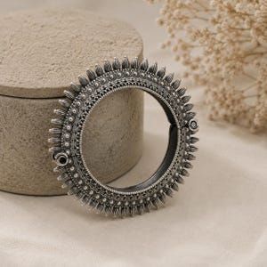 May include: A circular silver-toned bangle bracelet featuring an oxidised finish with intricate embossed patterns and a radiating sunburst edge design. The bangle includes a hinged opening mechanism with a small circular clasp on the side.