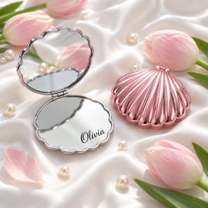 Op de afbeelding: Een schelpvormige compacte spiegel in roségoud met een scharnierend ontwerp. Eén kant heeft een reflecterend oppervlak met de naam Olivia in zwarte schrijfletters, terwijl de andere kant een gewone spiegel is. Een gesloten bijpassende spiegel.