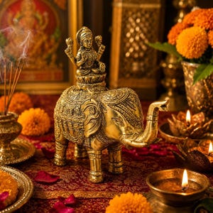 May include: A metallic gold-toned elephant figurine featuring intricate embossed patterns across its body and back. A small deity figure sits atop the elephant on a lotus base, holding lotus flowers in its hands. The sculpture has a polished finish.