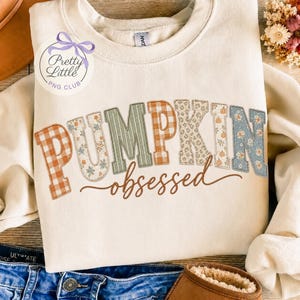 May include: A cream-colored crewneck sweatshirt featuring the word PUMPKIN in large, block-style letters with varied patterns including orange gingham, floral prints, green stripes, and leopard spots, with the word obsessed written in brown script.