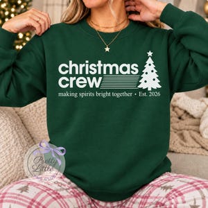 May include: A dark green long-sleeve crewneck sweatshirt featuring white text that reads "christmas crew" with a stylized Christmas tree graphic and the additional text "making spirits bright together &bull; Est. 2026" printed on the front.