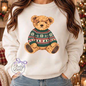 May include: A white crewneck sweatshirt featuring a central graphic of a brown teddy bear wearing a green, red, and white patterned jumper with snowflake and tree motifs. The sweatshirt has long sleeves and a round neckline.