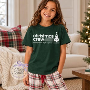 May include: A dark green short-sleeved t-shirt featuring white text that reads "christmas crew" with horizontal lines, a graphic of a Christmas tree, and the text "making spirits bright together &bull; Est. 2026" printed on the front.