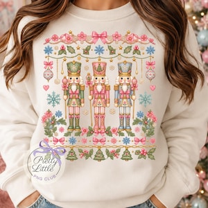 May include: A cream-colored sweatshirt featuring a graphic print of three nutcrackers in green, pink, and blue uniforms. The design includes hanging ornaments, bows, snowflakes, holly, and small Christmas trees. The text Pretty Little PNG CLUB is visible.
