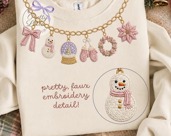 May include: A cream-coloured sweatshirt featuring a gold chain graphic with hanging charms including a pink bow, a snowman, a snow globe, pink mittens, a wreath, and a poinsettia. Text reads: pretty, faux embroidery detail! and a snowman.