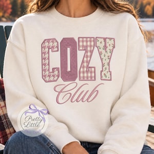 May include: A cream-colored crewneck sweatshirt featuring the word COZY in large block letters with varied patterns including houndstooth, solid mauve, gingham, and a floral print. Below, the word Club is written in a cursive mauve script font.
