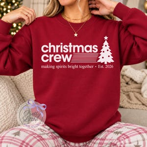 May include: A dark red long-sleeve sweatshirt featuring white text that reads "christmas crew" with a graphic of a stylized Christmas tree, followed by the text "making spirits bright together &bull; Est. 2026" printed on the front.