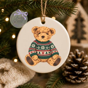 May include: A round white ceramic ornament featuring a brown teddy bear wearing a green, red, and white patterned holiday jumper with snowflake and tree motifs. The ornament hangs from a jute twine loop and includes a purple logo graphic.