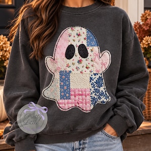 May include: A charcoal gray crewneck sweatshirt featuring a large ghost-shaped applique on the chest. The ghost is constructed from various patchwork fabric squares in shades of pink, blue, and white, featuring floral, lace, and woven patterns.