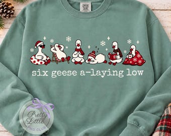 May include: A sage green crewneck sweatshirt featuring a graphic of six white geese in various relaxed poses, including wearing a Santa hat, reading, and wrapped in a blanket. The text below reads six geese a-laying low in white.