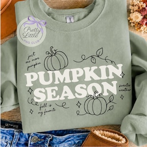 May include: A sage green crewneck sweatshirt featuring a graphic with two black line-art pumpkins, vines, and stars. The text reads: "just one more pumpkin", "PUMPKIN SEASON", "sweater weather", and "fall is my favorite". A circular logo is visible.