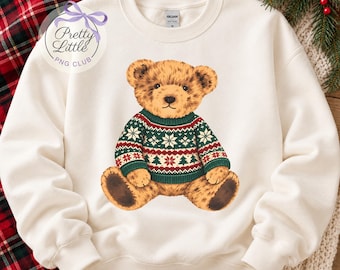 May include: A cream-colored crewneck sweatshirt featuring a graphic of a brown teddy bear wearing a green, red, and white patterned sweater with snowflake and tree motifs. The sweatshirt has a Gildan brand label at the inner neckline.