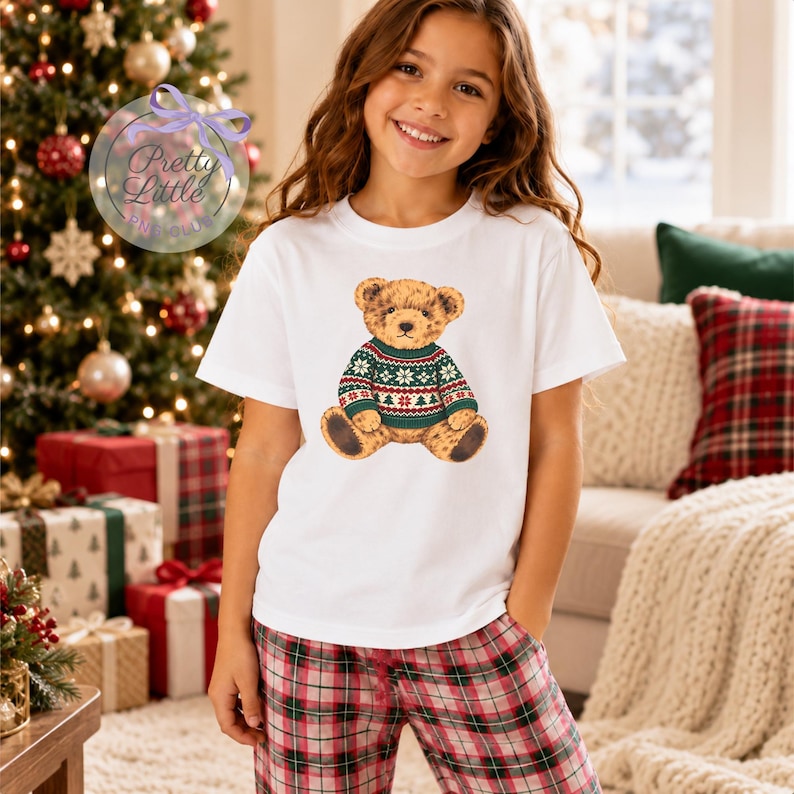 May include: A white short-sleeved t-shirt featuring a graphic of a brown teddy bear wearing a green, red, and white patterned holiday jumper. The bear is centred on the chest of the shirt. The shirt is paired with red and black plaid trousers.