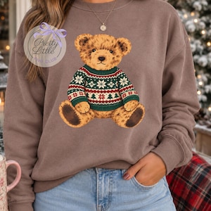 May include: A brown crewneck sweatshirt featuring a graphic of a teddy bear wearing a green, red, and white patterned holiday sweater with snowflakes and trees. The graphic includes a circular logo with the text Pretty Little PNG CLUB.