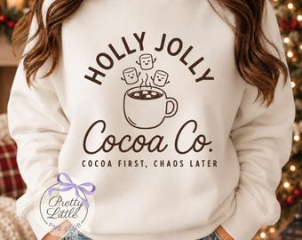 May include: A white crewneck sweatshirt featuring a brown graphic of a mug of hot cocoa with three smiling marshmallows above it. The text reads HOLLY JOLLY Cocoa Co. and COCOA FIRST, CHAOS LATER in a dark brown font on the front center.