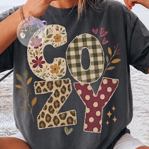 May include: A charcoal gray short-sleeve t-shirt featuring the word COZY in large letters. The C has a floral pattern, the O has a green gingham check, the Z has a leopard print, and the Y has a maroon polka dot pattern with small heart icons.