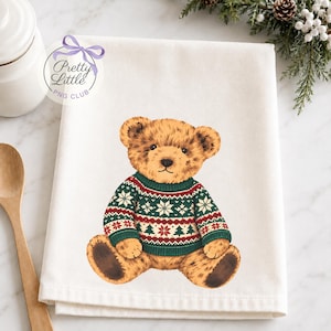 May include: A white kitchen towel featuring a centred illustration of a brown teddy bear wearing a green, red, and white knit jumper with a snowflake and pine tree pattern. The bear is sitting down with dark brown paw pads.