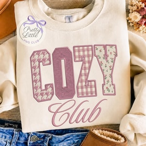 May include: A cream-colored crewneck sweatshirt featuring the text COZY CLUB in large varsity-style letters. The letters are filled with various patterns including houndstooth, solid mauve, gingham, and a small floral print in shades of pink and white.