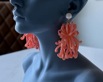 May include: A matte dark gray bust of a person with hair styled in a bun, wearing a pair of dangling earrings. The earrings feature a small white spherical stud at the top with a cluster of thin, coral-colored, curved plastic strands hanging below.