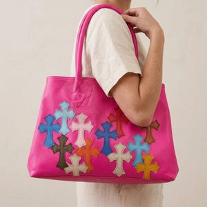 Puede incluir: Un bolso tote de color rosa brillante con una superficie texturizada y dos correas para el hombro. El exterior está decorado con múltiples apliques en forma de cruz de varios colores, incluyendo azul claro, azul oscuro, rojo, amarillo, marrón y crema.