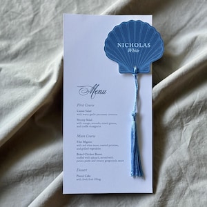 May include: A white rectangular menu card featuring the text "Menu," "First Course," "Main Course," and "Dessert" with specific food items listed. A blue seashell-shaped card with the text "NICHOLAS White" is attached by a blue tassel.