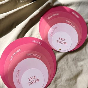 May include: Two circular menu sets featuring stacked paper layers in shades of pink. Each set displays the text Kyle Taylor, April 17, 2026, First Course, Main Course, and Dessert in a serif font. The layers are fastened together with a small gold stud.