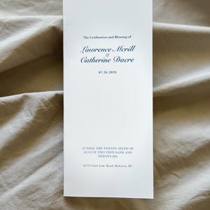 May include: A tall, rectangular white wedding programme card with dark blue text. It reads: The Celebration and Blessing of Lawrence Mcrill & Catherine Dacre, 07.26.2026, SUNDAY, THE TWENTY SIXTH OF AUGUST TWO THOUSAND AND TWENTY-SIX, 4870 Chute Lake Road, Kelowna, BC.