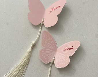 Könnte beinhalten: Zwei schmetterlingsförmige Lesezeichen aus Papier in Hellrosa. Ein Lesezeichen trägt den Namen James in dunkelroter Schreibschrift, das andere den Namen Sarah in Dunkelrot mit weißem Flügelmuster. Beide haben eine lange, cremefarbene Quaste.
