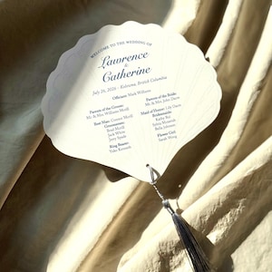 Peut inclure: Un éventail de programme de mariage en forme de coquillage en papier cartonné blanc cassé avec des bords festonnés. Le texte indique : Welcome to the Wedding of Lawrence & Catherine, July 26, 2026, Kelowna, British Columbia, avec un pompon gris.