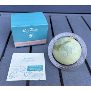 Könnte beinhalten: Ein Max Factor World of Beauty Lotion Set bestehend aus einer türkisfarbenen Pappschachtel, einem kugelförmigen hellgrünen Lotionsbehälter mit einem durchsichtigen Ring mit der Aufschrift World of Beauty Lotion und einem Beipackzettel.