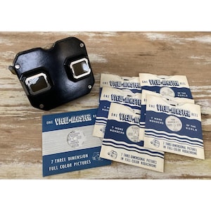 Puede incluir: Un visor estereoscópico View-Master de plástico negro con dos lentes rectangulares, acompañado de varias fundas de papel etiquetadas como View-Master Reel, cada una con siete imágenes tridimensionales a todo color en formato Kodachrome.