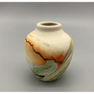 May include: A small, rounded ceramic vase with a matte, off-white base color. The surface features swirling, marbled patterns in shades of burnt orange, sage green, and dark brown, creating a fluid, organic design around the exterior body.