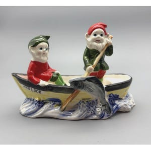 May include: A ceramic figurine featuring two gnomes in a yellow rowboat with the text GOOD LUCK printed on the side. One gnome wears a red outfit and green hat, while the other wears a green outfit and red hat, holding an oar with a grey fish.